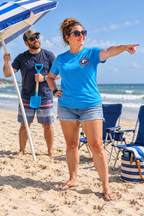 Illustrative lifestyle mockup: white woman in a turquoise OVE tee directing placement of a beach umbrella.
